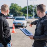 A male driving instructor provides guidance to a male student as they both look at a car